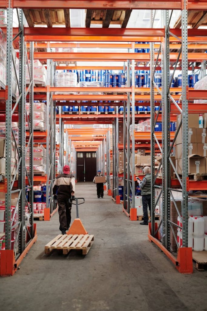 men-working-in-factory-warehouse-4483772 Workers arrange inventory on shelves in a large industrial warehouse.