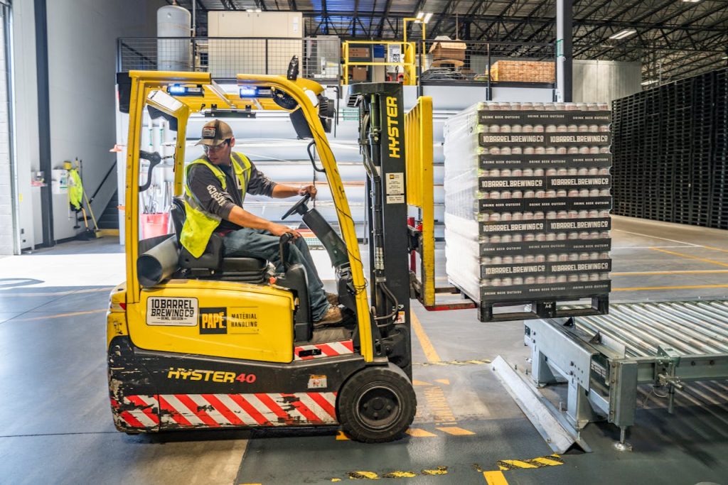person-using-forklift-1267338 A warehouse worker maneuvers a forklift to transport crates for brewing company storage.